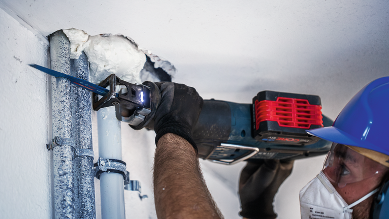 A person wearing safety equipment cuts through a ceiling pipe using a reciprocating saw.