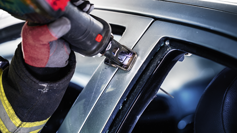 Person wearing safety equipment uses a power tool to cut through a car frame.