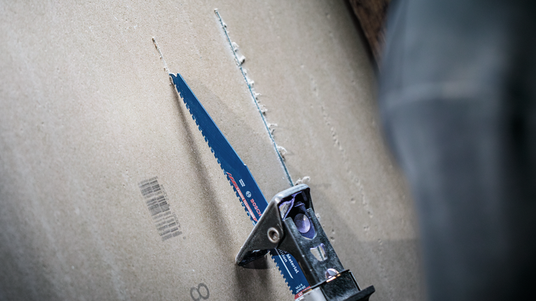 A person wearing safety equipment uses a reciprocating saw to cut drywall.