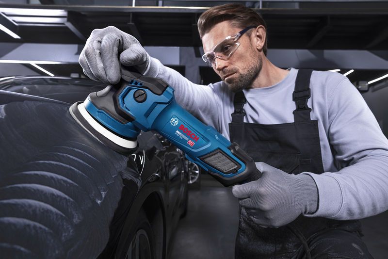 A person wearing safety equipment polishes a black car with an electric polisher.