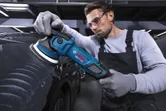 A person wearing safety equipment polishes a black car with an electric polisher.