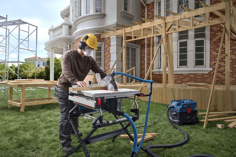 A person wearing safety equipment cuts wood with a table saw at a house construction site.