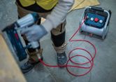 A person wearing safety equipment uses a pneumatic nail gun connected to an air compressor.