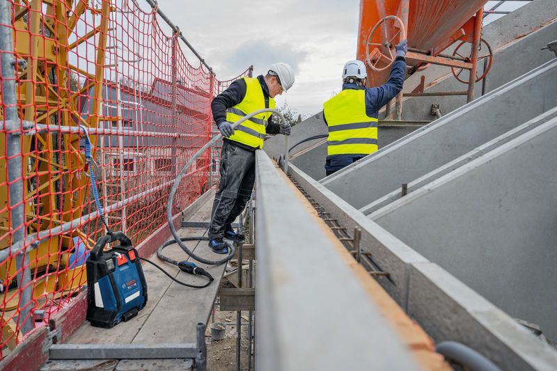 Two persons wearing safety equipment work on scaffolding with construction tools and concrete forms.
