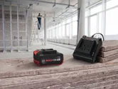 Rechargeable battery and charger on a wooden surface, with a person working on a ladder in the background.
