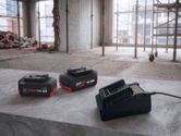 Two rechargeable tool batteries and a charger on a concrete surface in a construction area.