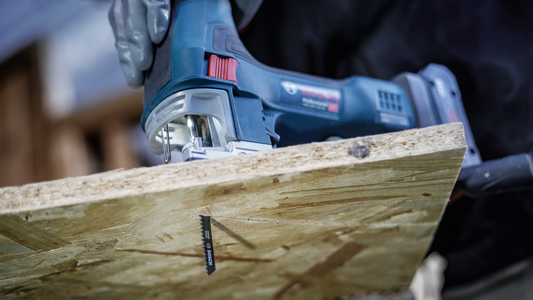 Person wearing safety equipment cuts a wood board with a cordless jigsaw.