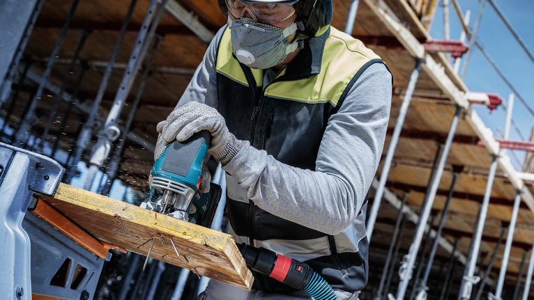 Person wearing safety equipment cuts wood with a jigsaw at a construction site.