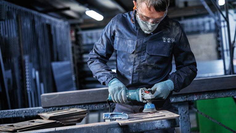A person wearing safety equipment uses a power tool to grind metal.