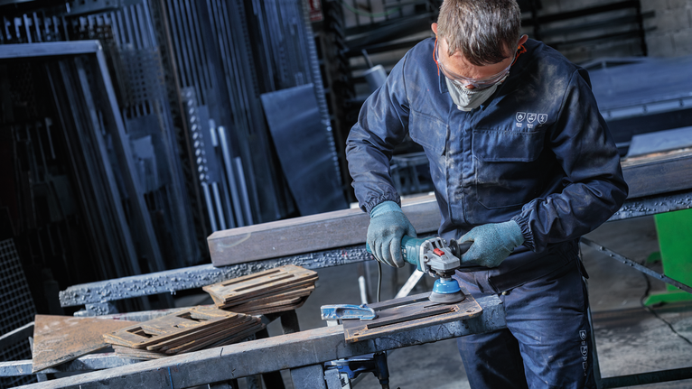 Person wearing safety equipment grinds metal parts on a workbench.