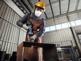 A person wearing safety equipment uses a cordless straight grinder on a metal beam.