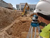 Person wearing safety equipment uses an optical level at a construction site.