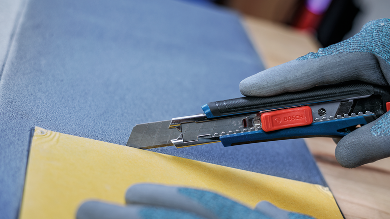 Person wearing safety equipment cuts yellow paperboard with a utility knife.