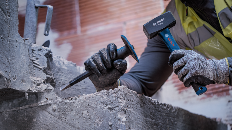 Person wearing safety equipment uses a hammer and chisel to break concrete.