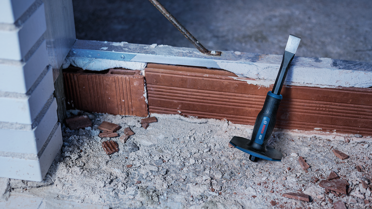 Chisel tool placed on dusty construction site floor near a partially demolished brick wall.