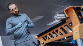 Person wearing safety equipment inspects two metal tools beside an orange industrial frame.