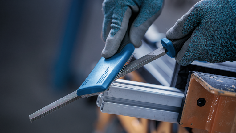 Person wearing safety equipment files a metal piece clamped in a vise.