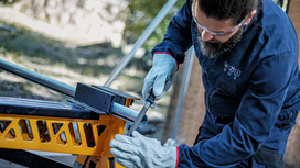 Person wearing safety equipment files a metal pipe on a yellow workbench.