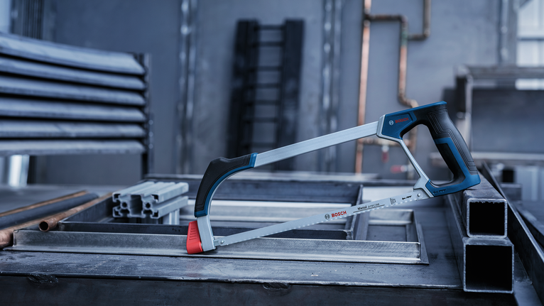 A metal hand saw rests on stacked steel beams in an industrial workshop.