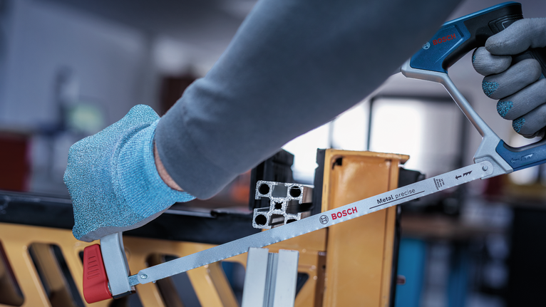 Person wearing safety equipment cuts a metal bar with a hacksaw.