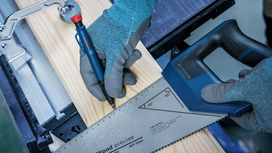 Person wearing safety equipment measures and marks a wooden board with a saw and pencil.