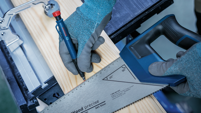 Person wearing safety equipment measures and marks a wooden board with a saw and pencil.