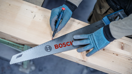 Person wearing safety equipment marks a wooden plank with a saw and pencil.