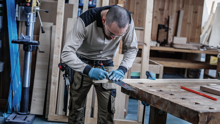 Person wearing safety equipment measures a wooden plank in a workshop.