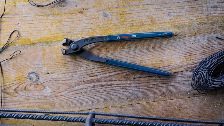 Steel nipper pliers lie on a wooden workbench beside coiled wire and rebar.