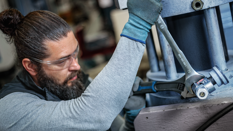 A person wearing safety equipment tightens a large bolt with a wrench on industrial machinery.