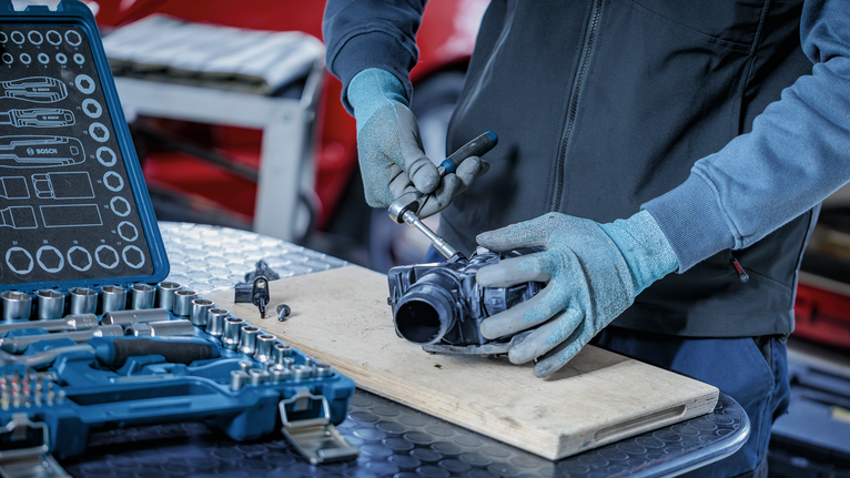 Person wearing safety equipment uses a ratchet tool to repair a mechanical part.