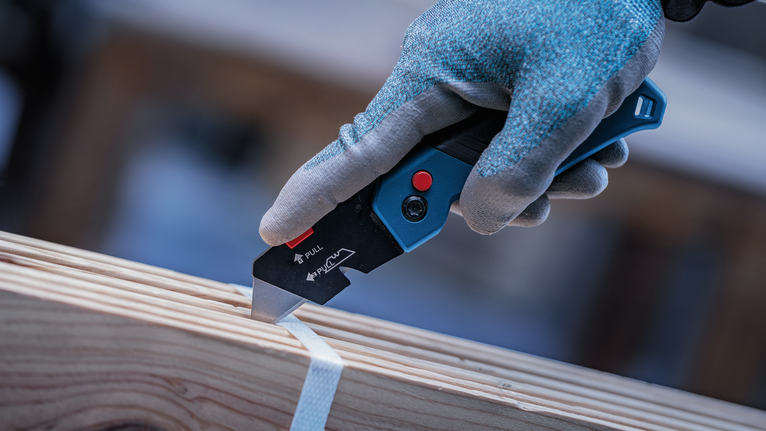 Person wearing safety equipment uses a utility knife to cut strapping on wood.