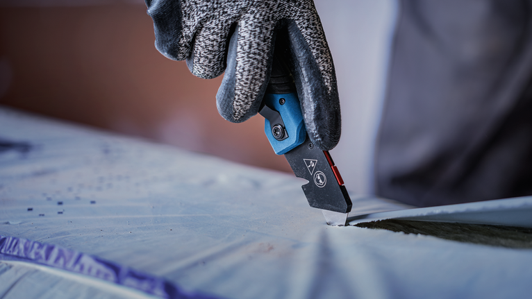 Person wearing safety equipment cuts plastic sheet with a utility knife.