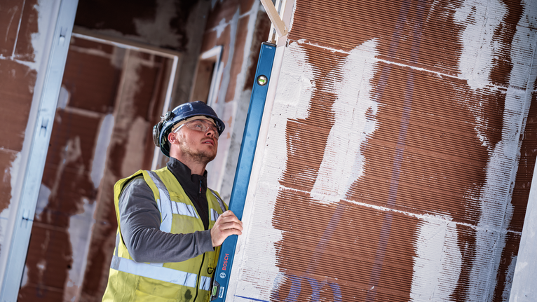 Person wearing safety equipment checks wall alignment with a spirit level.
