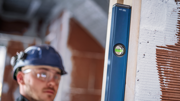 Person wearing safety equipment levels a vertical wooden beam on a construction site.