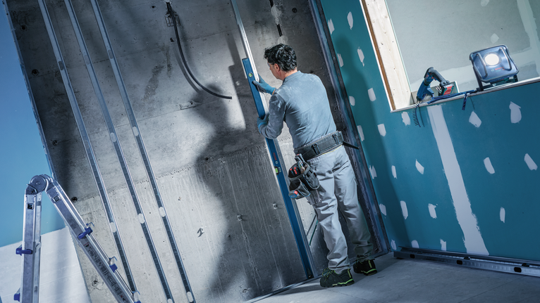 Person wearing safety equipment uses a spirit level to check alignment on a concrete wall.