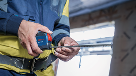 Person wearing safety equipment uses a measuring tape at a construction site.