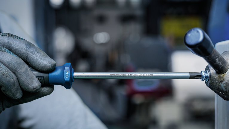 Person wearing safety equipment tightens a screw with a long screwdriver.