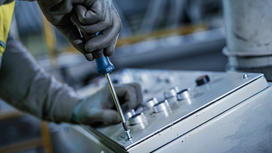 Person wearing safety equipment uses a screwdriver to fasten a panel on control equipment.
