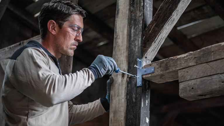 Person wearing safety equipment fastens a screw into a wooden beam with a power tool.