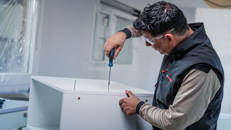 Person wearing safety equipment assembles white furniture using a screwdriver.