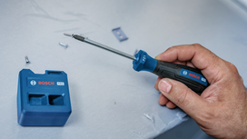 Person holding a screwdriver near a screw and a blue tool holder on a work surface.