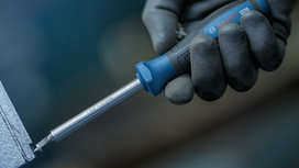 Person wearing safety equipment drives a screwdriver into a metal surface.