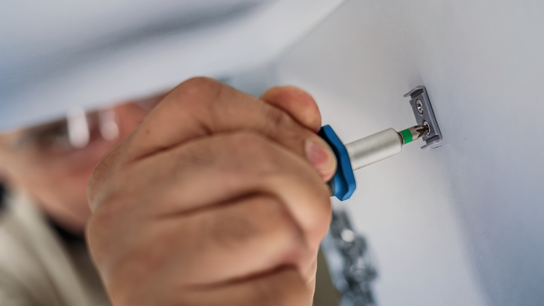 Person wearing safety equipment secures a metal bracket to a wall with a screwdriver.