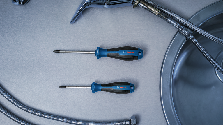Two screwdrivers placed beside flexible metal hoses and a kitchen sink.