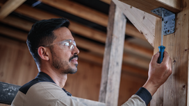 Person wearing safety equipment fastens a metal bracket to a wooden beam with a screwdriver.