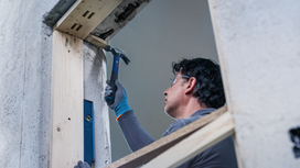 Person wearing safety equipment uses a hammer to work on a wooden window frame.