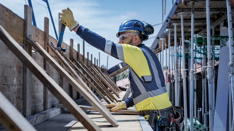 Person wearing safety equipment uses a hammer to secure wood at a construction site.