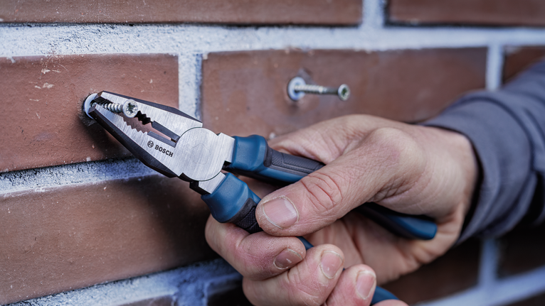 Person uses pliers to grip a screw inserted in a brick wall.