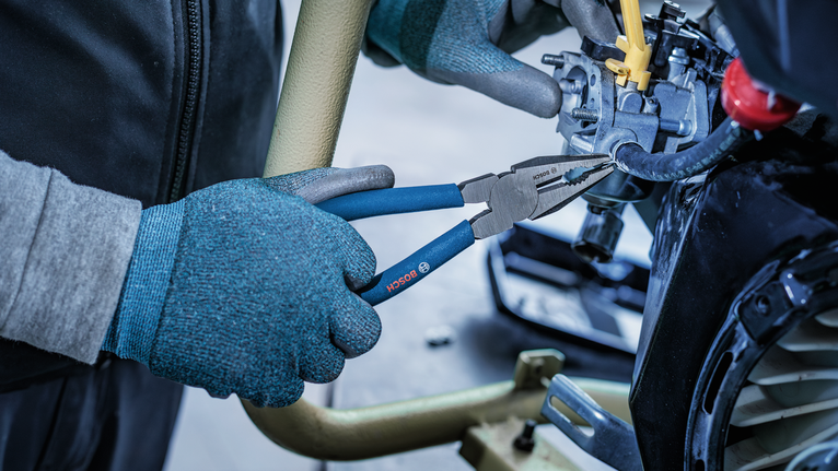 Person wearing safety equipment uses pliers to grip a small machine part.